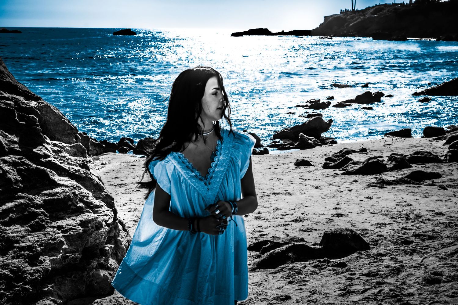Woman in blue dress standing on rocky beach with sparkling blue ocean behind her.