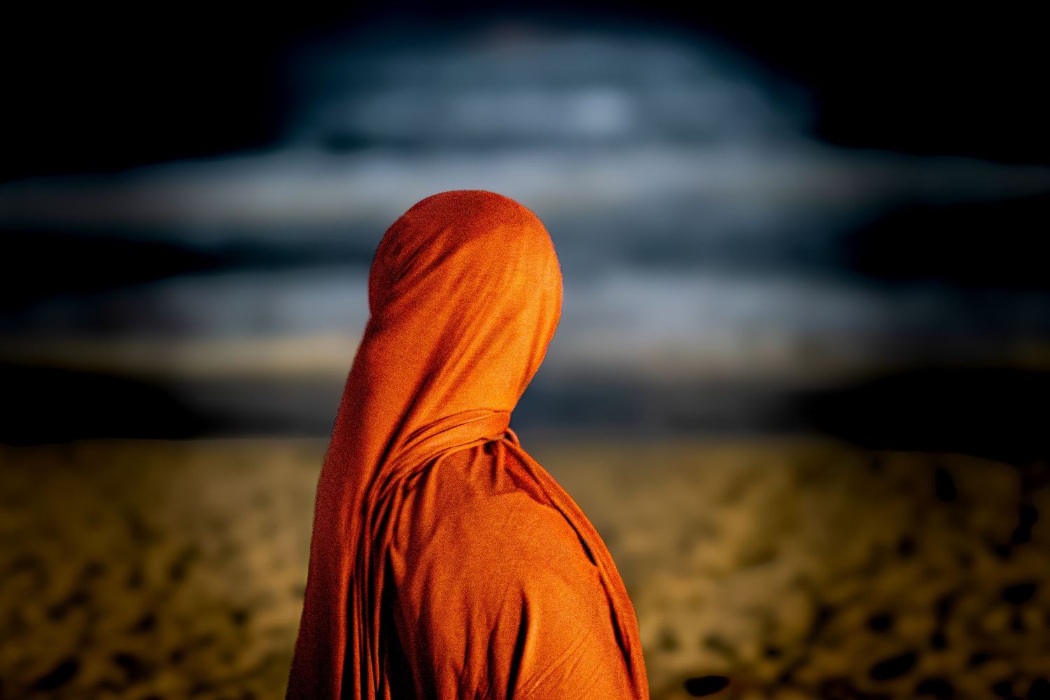 Person draped in orange fabric standing on a sandy beach at night with ocean waves in the background.