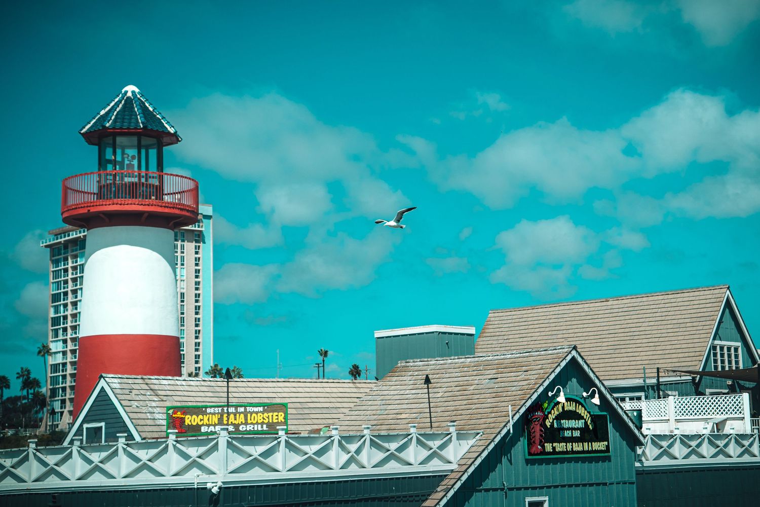 Red and white lighthouse tower rises above colorful seaside shops under a bright blue sky with a seagull in flight.