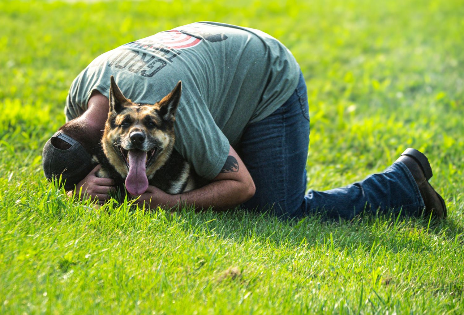 A man lying on green grass hugging a happy German Shepherd dog with tongue out.