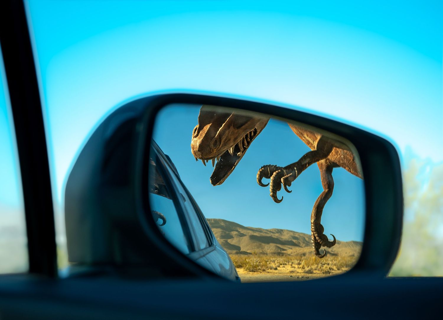 A car side mirror reflects a dinosaur statue in a desert landscape under a clear blue sky.