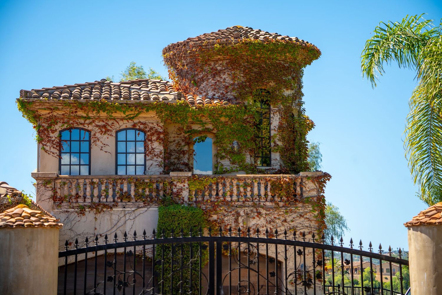 Ivy-covered Mediterranean villa with stone tower, arched windows, balcony, and wrought iron gate under clear blue sky.