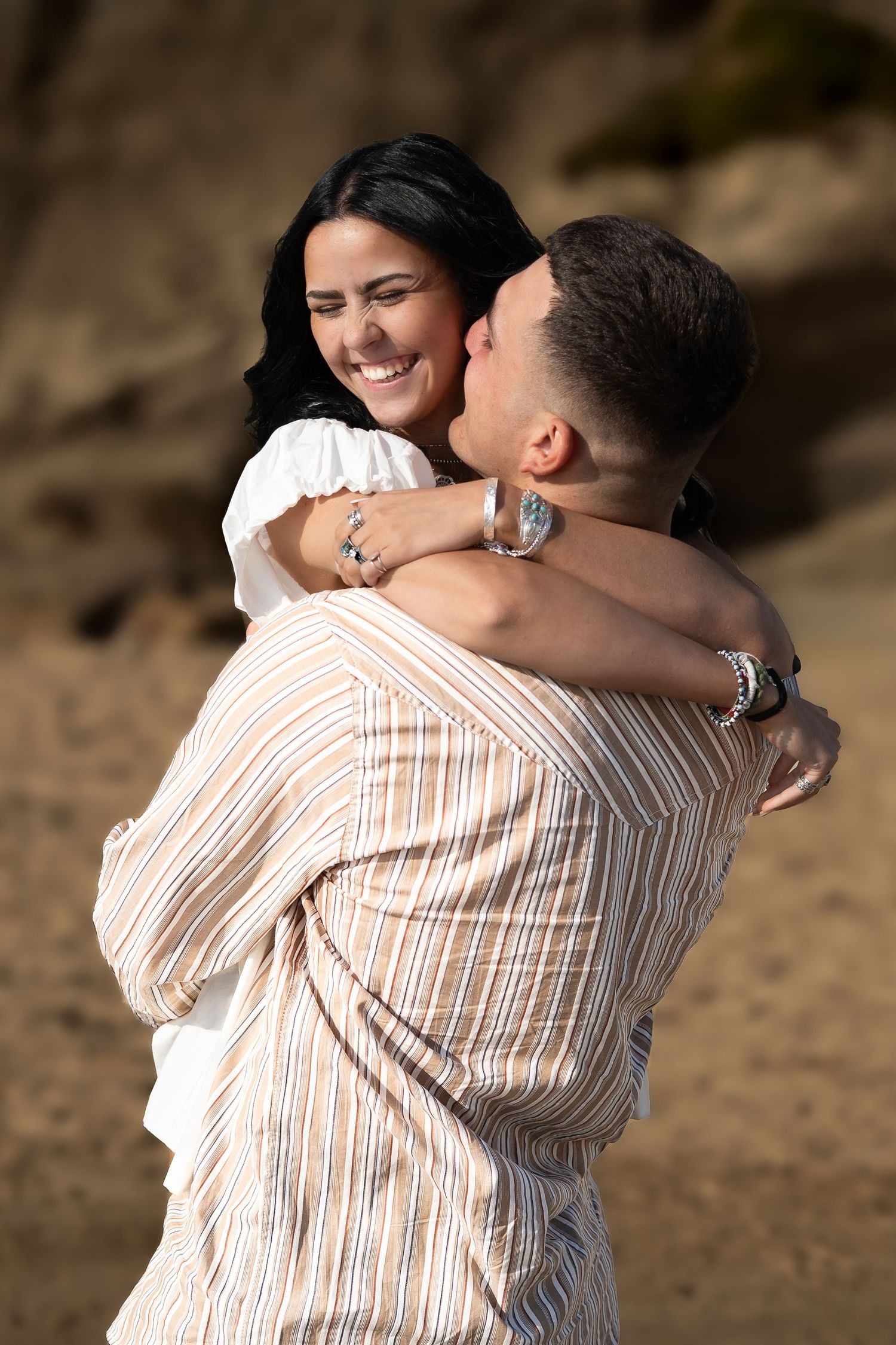 Smiling woman with dark hair embraces a man outdoors, wearing a white top and silver bracelet in warm sunlight.