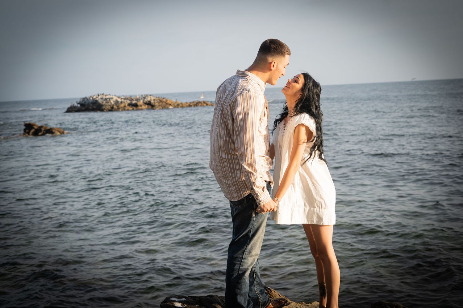 Romantic couple about to kiss while standing on rocks with calm ocean water behind them.