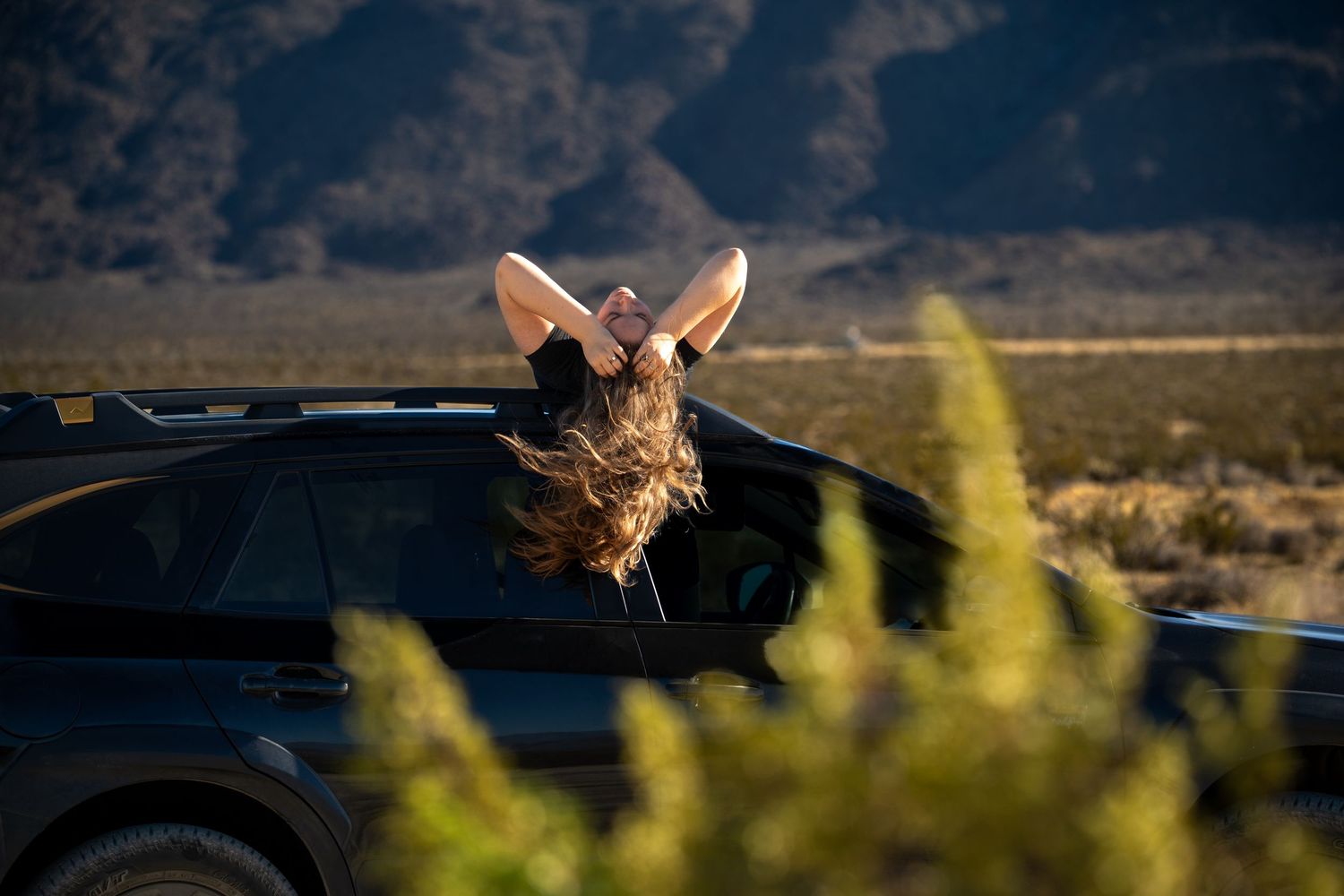 Person leaning out of a black car sunroof in a desert landscape with mountains in the background.