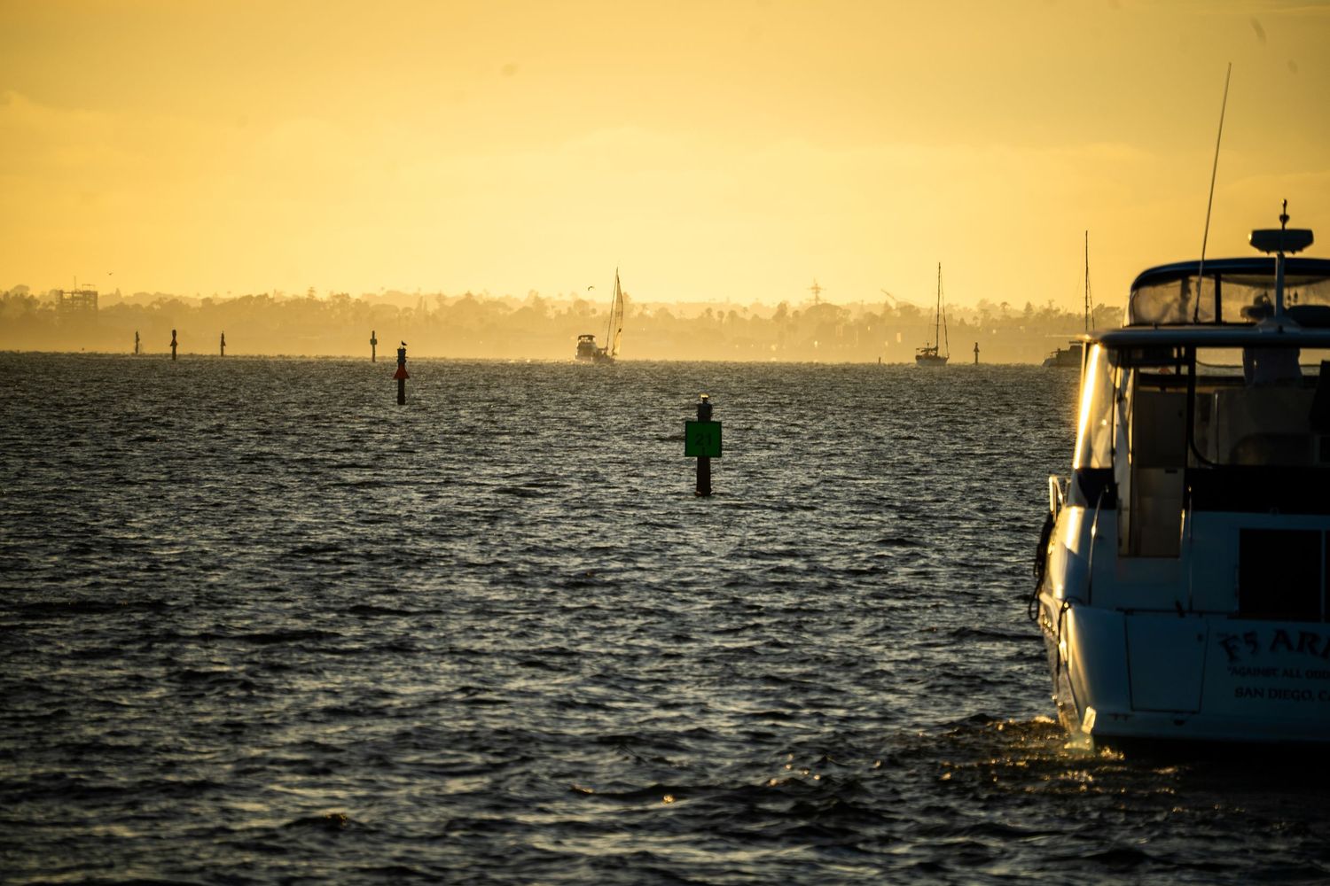 Golden sunset over a harbor with a green buoy, distant skyline, and a luxury yacht in the foreground on calm waters.