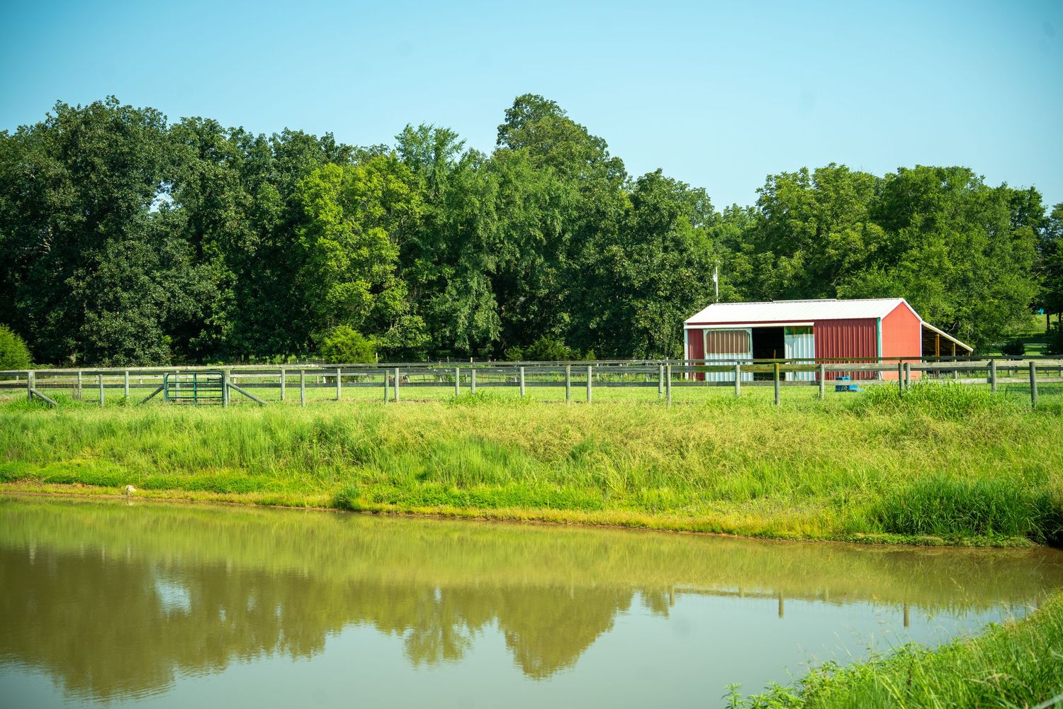 Red barn beside a wooden fence on a lush green farm with a calm reflective pond in the foreground.