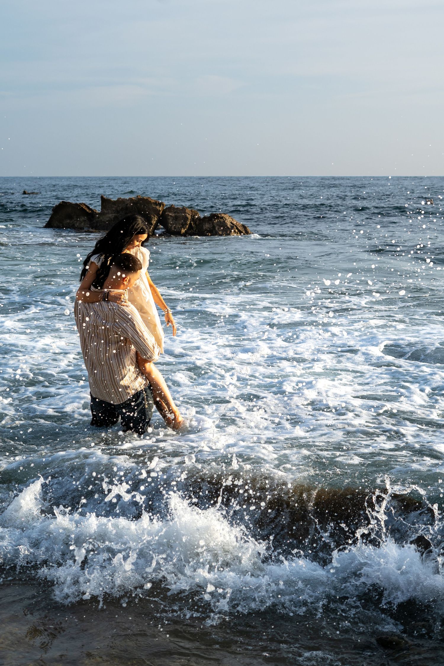 Couple embracing in ocean waves at rocky beach, woman in white dress, splashing water, sunny day.