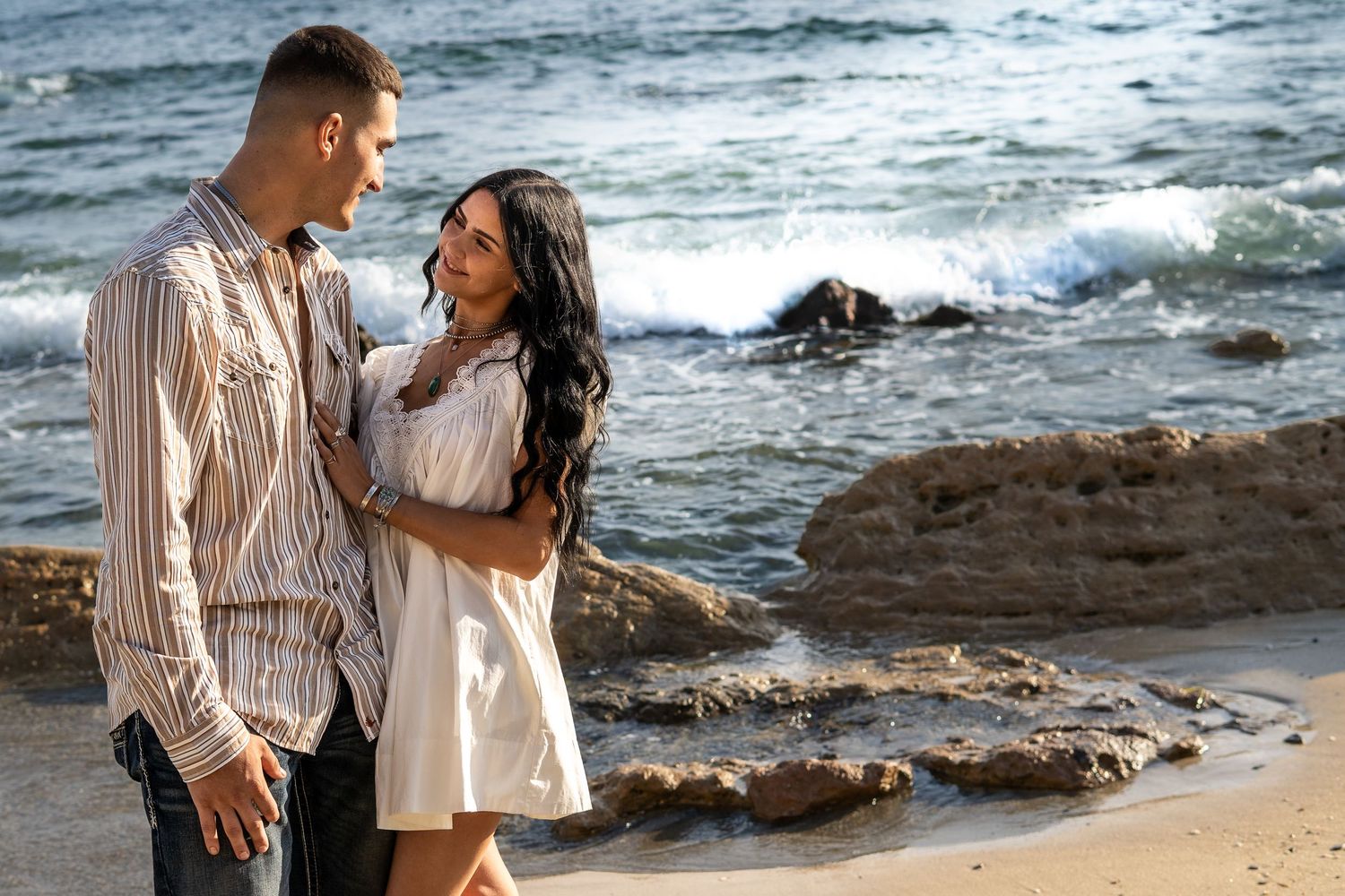 A romantic couple embraces on a rocky beach with ocean waves in the background.