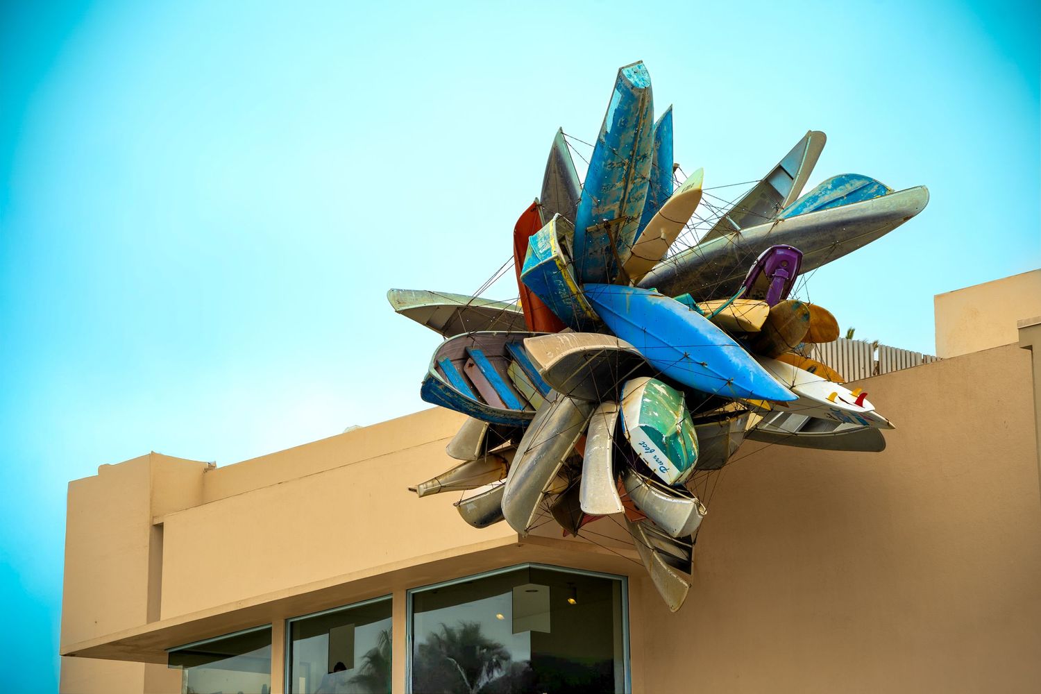 Colorful abstract metal sculpture made of surfboards mounted on top of a beige building against a blue sky.
