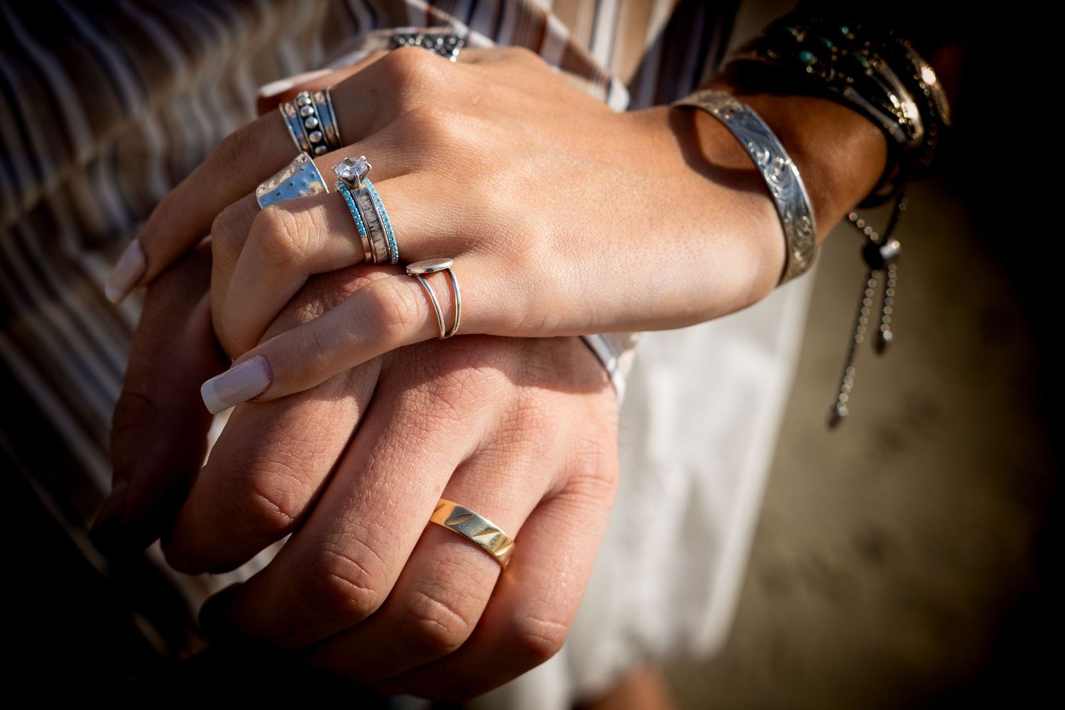 Two hands adorned with silver rings and bracelets, including turquoise stones, clasped together outdoors.