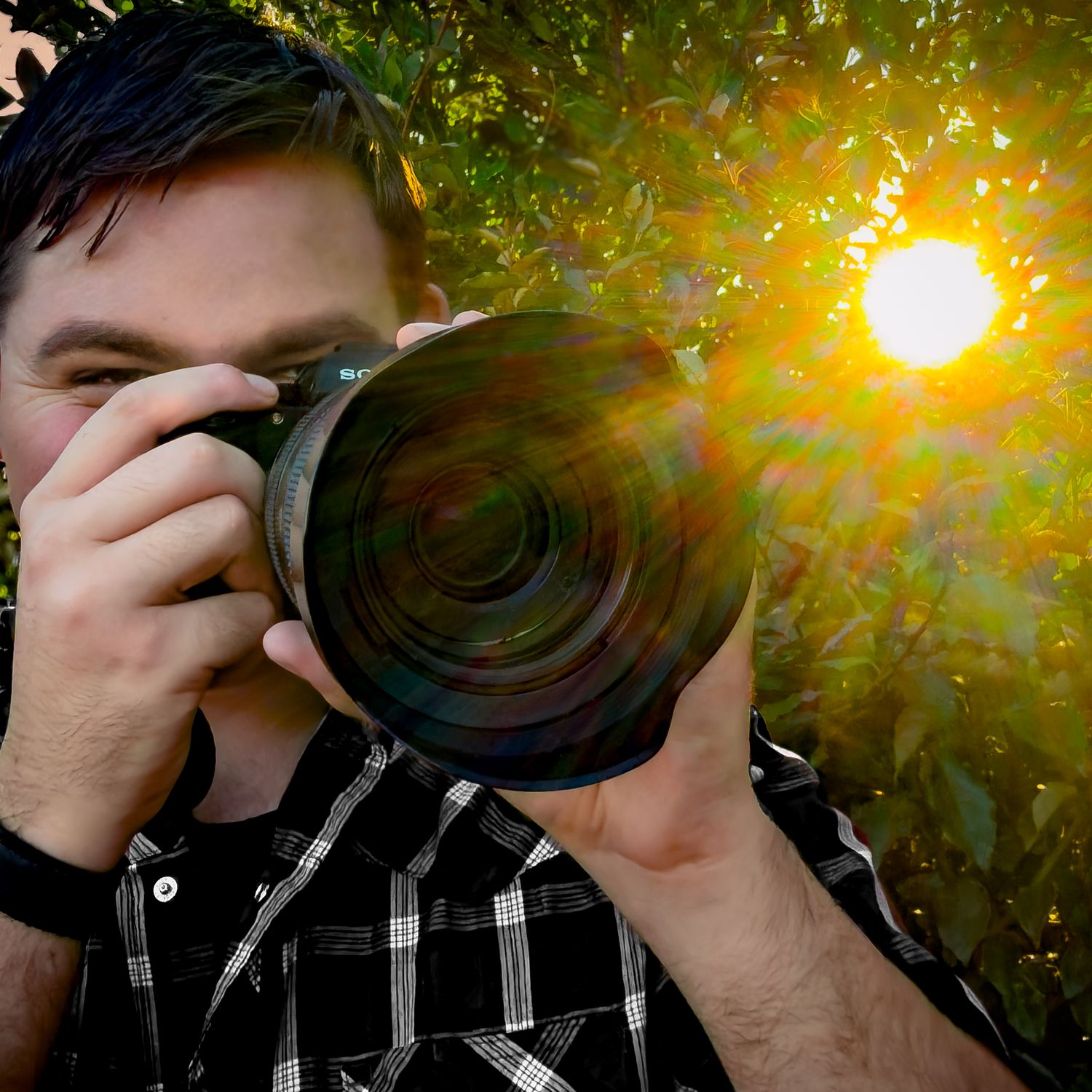 Man photographing with Sony camera outdoors, sun flare through green trees in background.