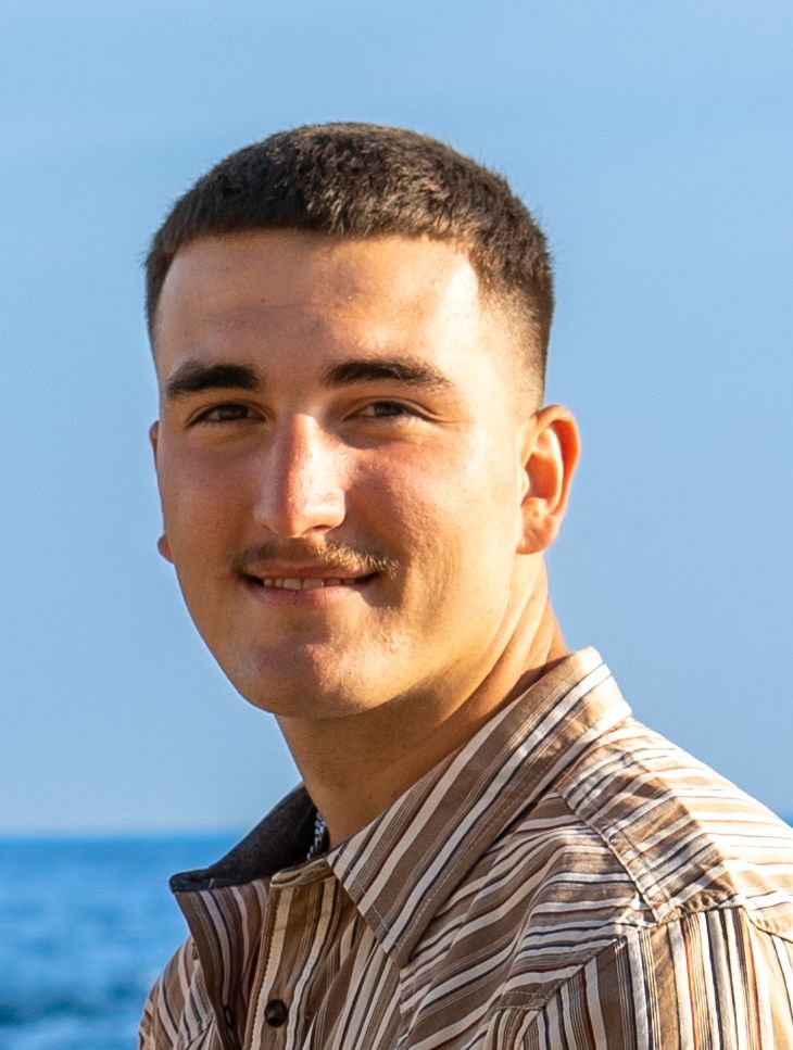 Young man with short dark hair smiling outdoors near the ocean on a sunny day, wearing a striped shirt.