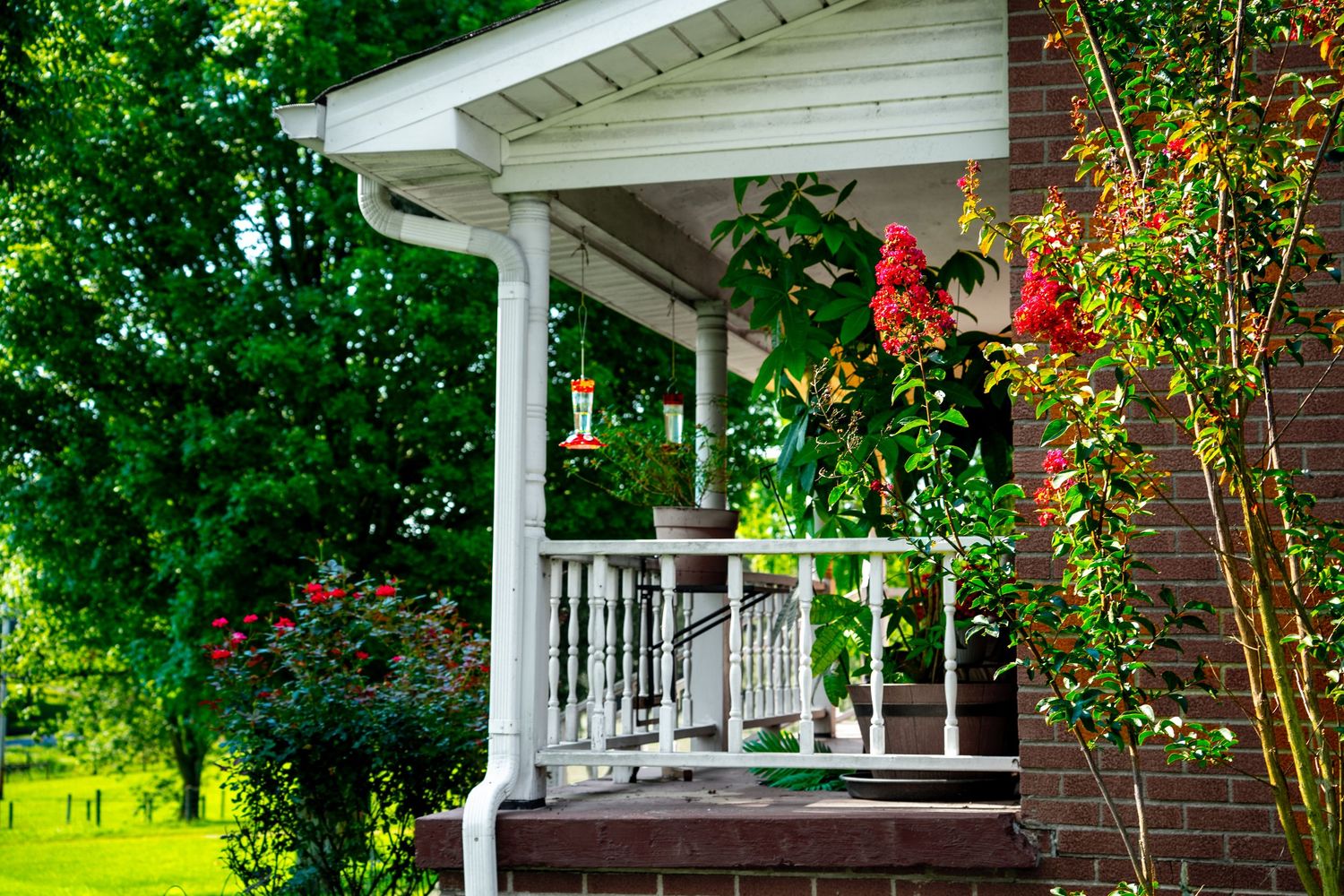 White porch with red flowers, hanging hummingbird feeder, and lush green trees surrounding a brick home.
