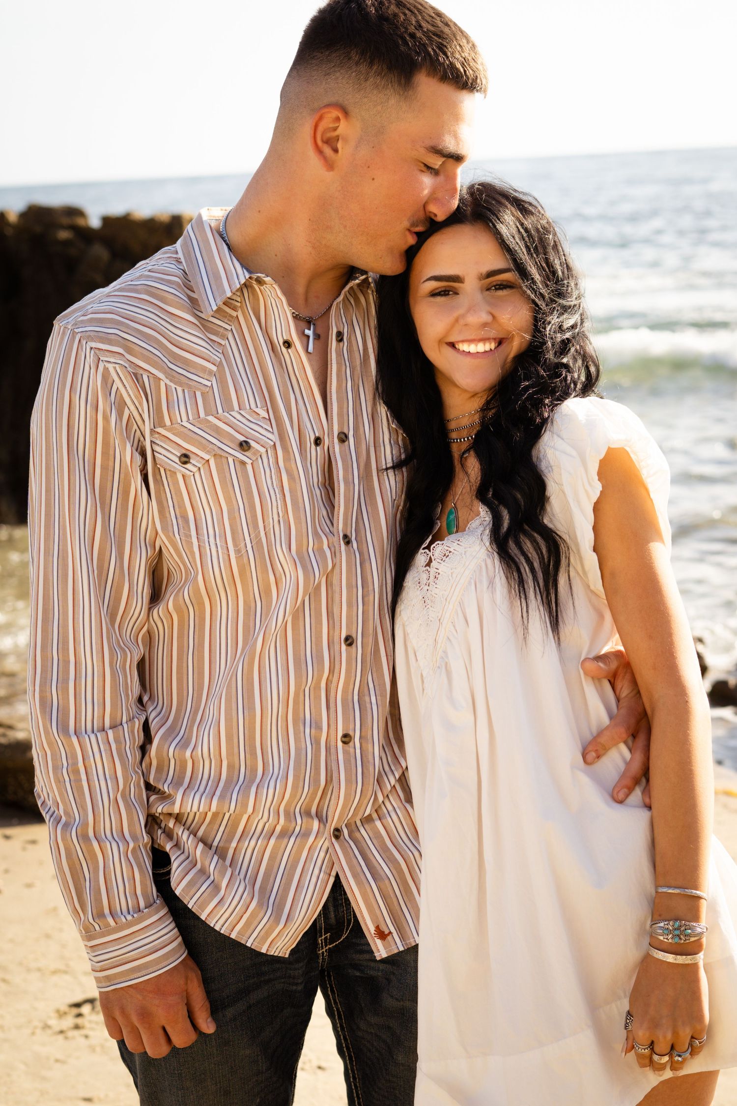 A couple embraces on a rocky beach, man in striped shirt kissing woman's head as she smiles in white dress.