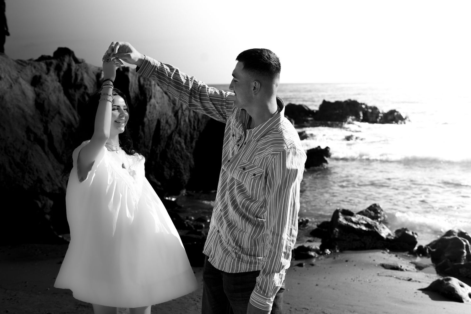 Black and white photo of a couple dancing on a rocky beach, man spinning woman in white dress.