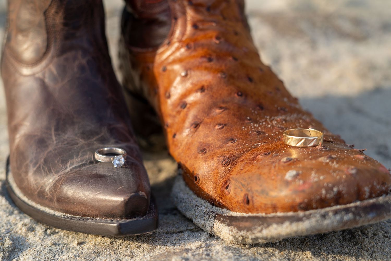 Two cowboy boots with wedding rings resting on them, one brown and one orange leather boot.