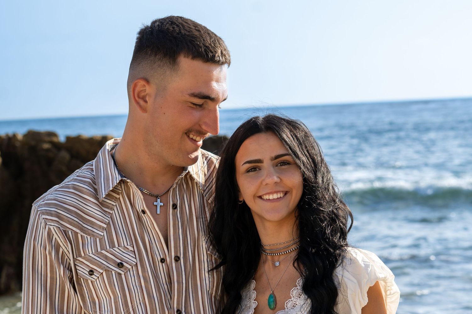 A smiling couple poses together on a sunny beach with ocean waves and rocky cliffs in the background.