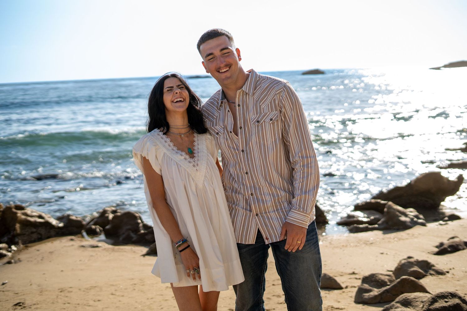 Smiling couple posing together on a sunny beach with ocean waves and rocks in the background.