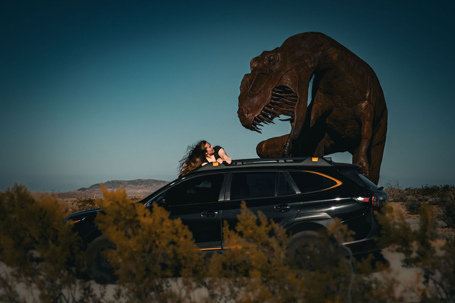Person coming up from SUV sunroof beside a large T-Rex dinosaur sculpture in a desert landscape. Dramatic, Jurassic inspired image.