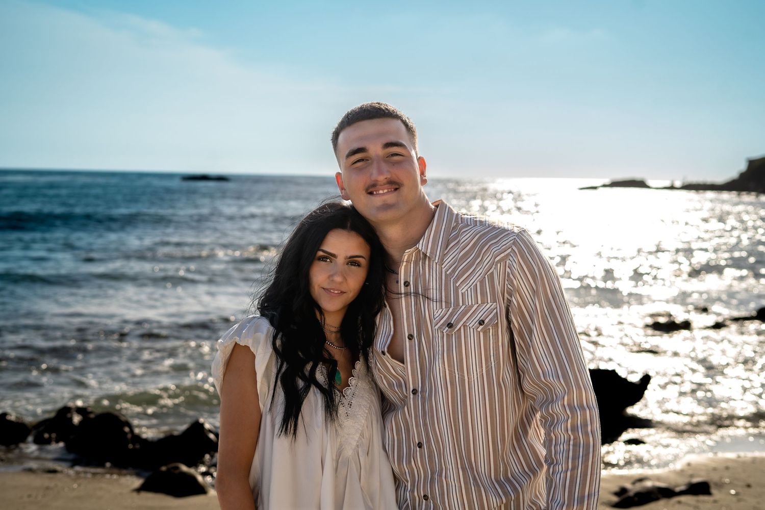 Happy couple smiling together on a sunny beach with sparkling ocean water in the background.