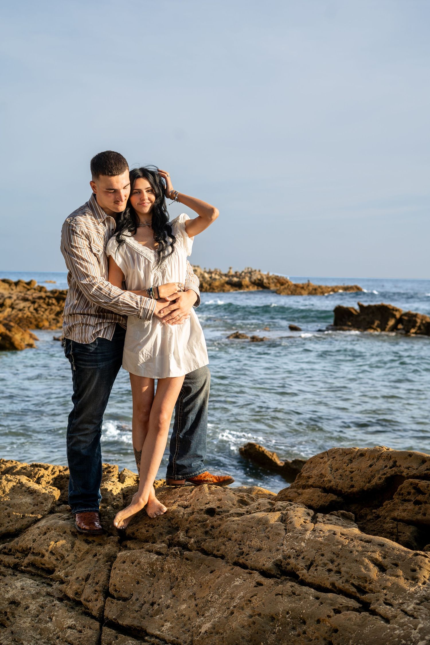 Romantic couple hugging on coastal rocks by the ocean, woman in white dress with hand in hair.