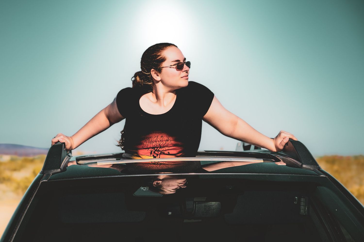 Woman in sunglasses leaning out of a car sunroof on a sunny desert road trip adventure.