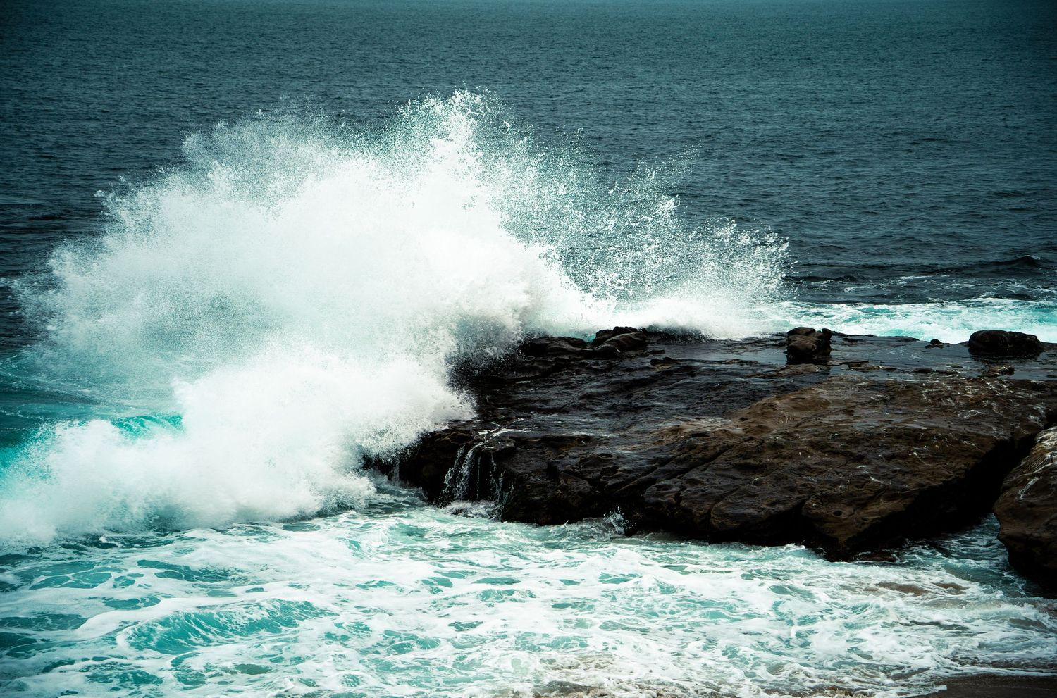 Ocean waves crashing dramatically against dark rocky coastline with white sea spray under a blue sky.
