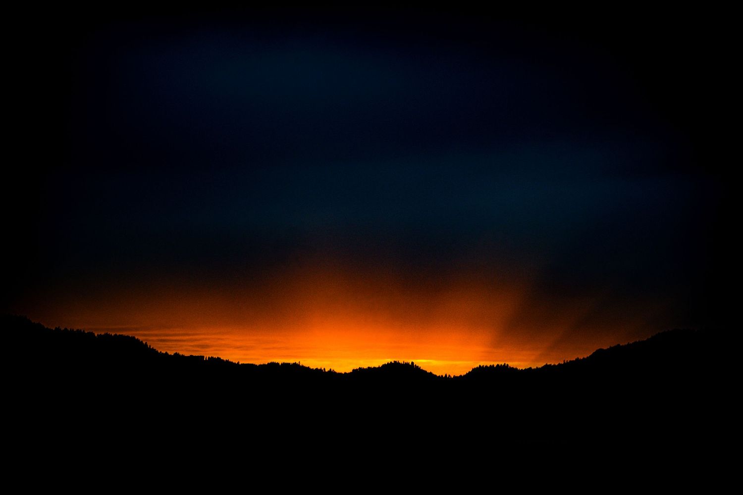 Dramatic night landscape with glowing orange horizon light silhouetting dark mountain ridges against a deep blue stormy sky.