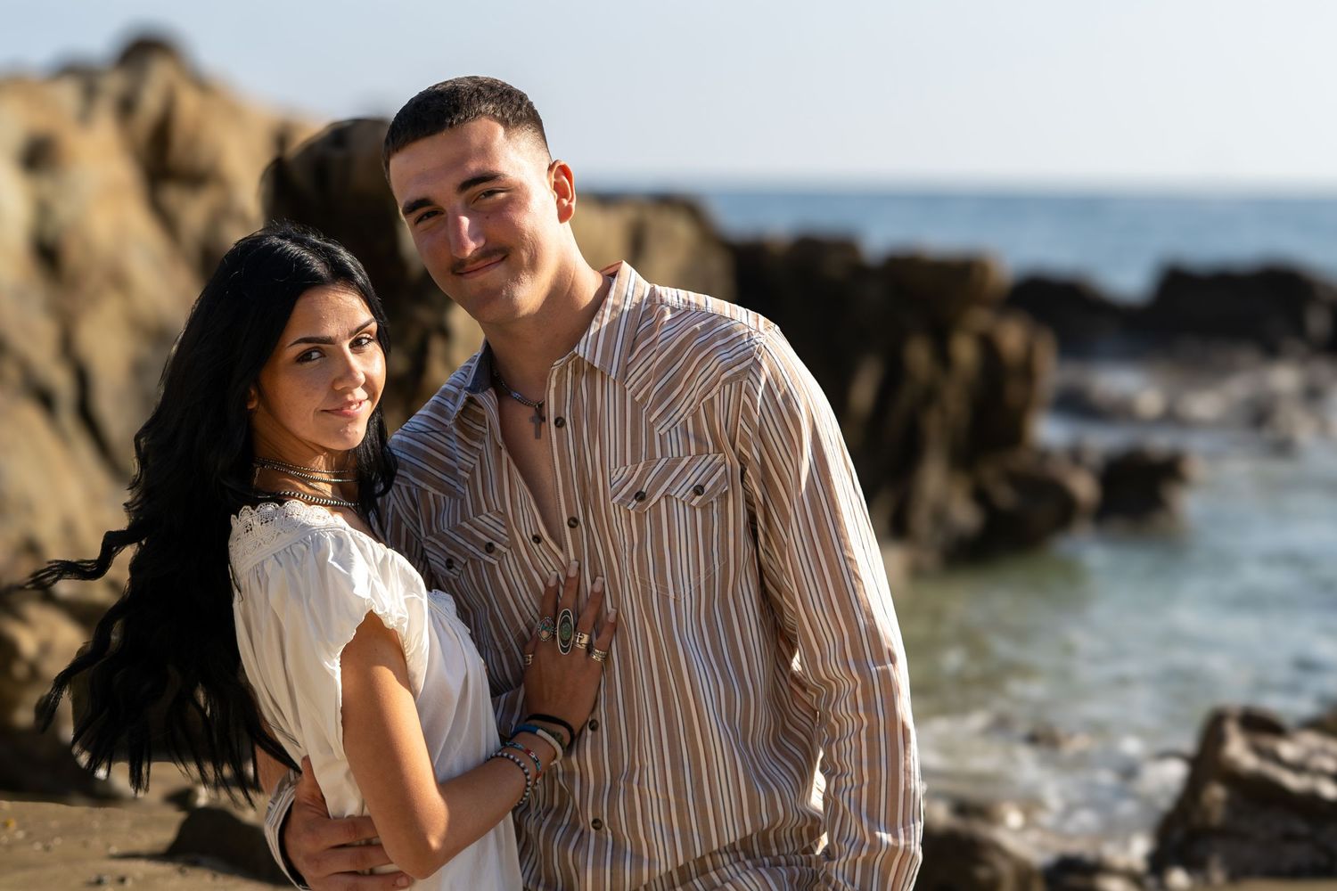 A young couple poses romantically on a rocky beach, the woman in white and man in striped shirt, with ocean waves behind them.