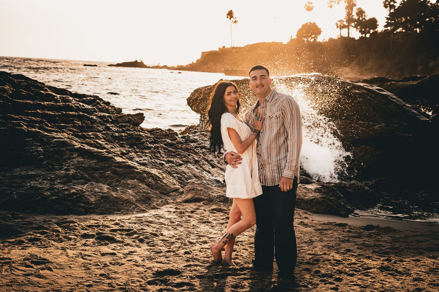 Happy couple posing on rocky coastline at golden sunset, woman in white dress, waves splashing.