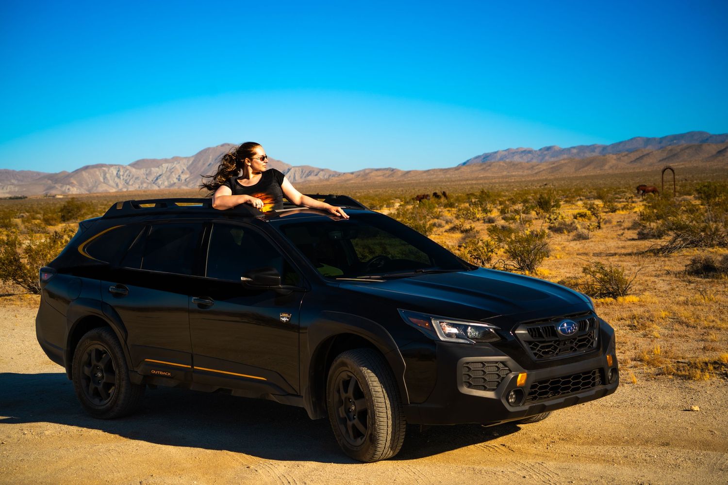Woman relaxing on roof of black Subaru Outback in vast desert landscape under clear blue sky.