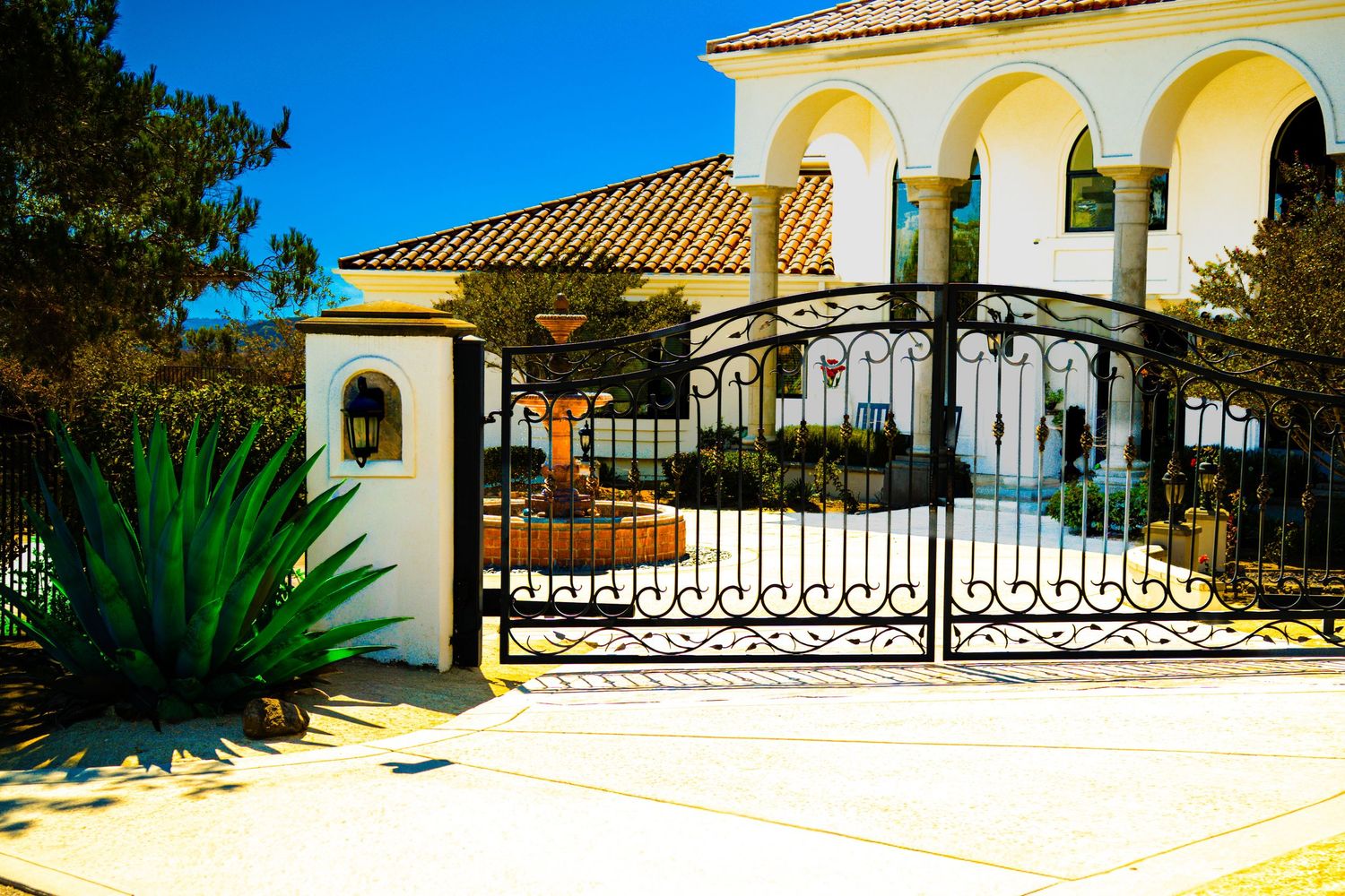 Elegant Mediterranean villa with ornate wrought iron gate, arched windows, terracotta roof, and lush agave plants in sunny landscape.