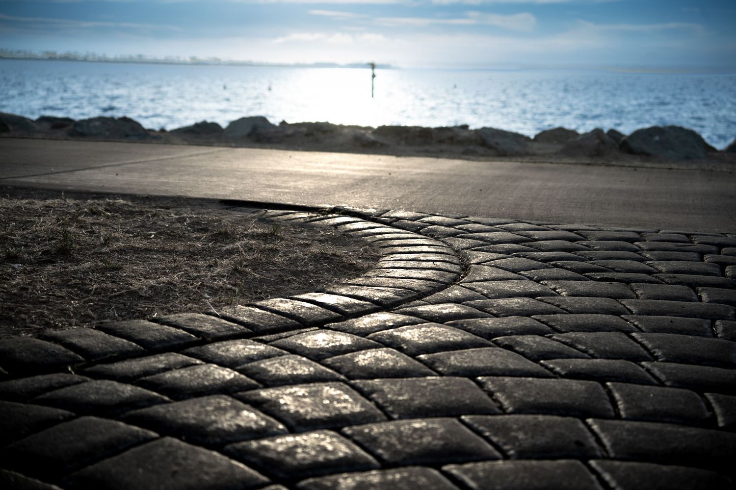 Curved cobblestone path leading to a rocky shoreline with calm ocean waters and a hazy horizon in soft natural light.