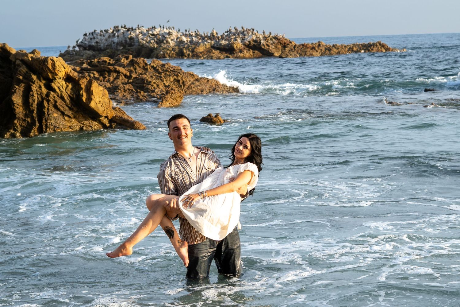 Couple enjoying the ocean, man carrying woman in his arms while standing in shallow coastal waters near rocky shore.