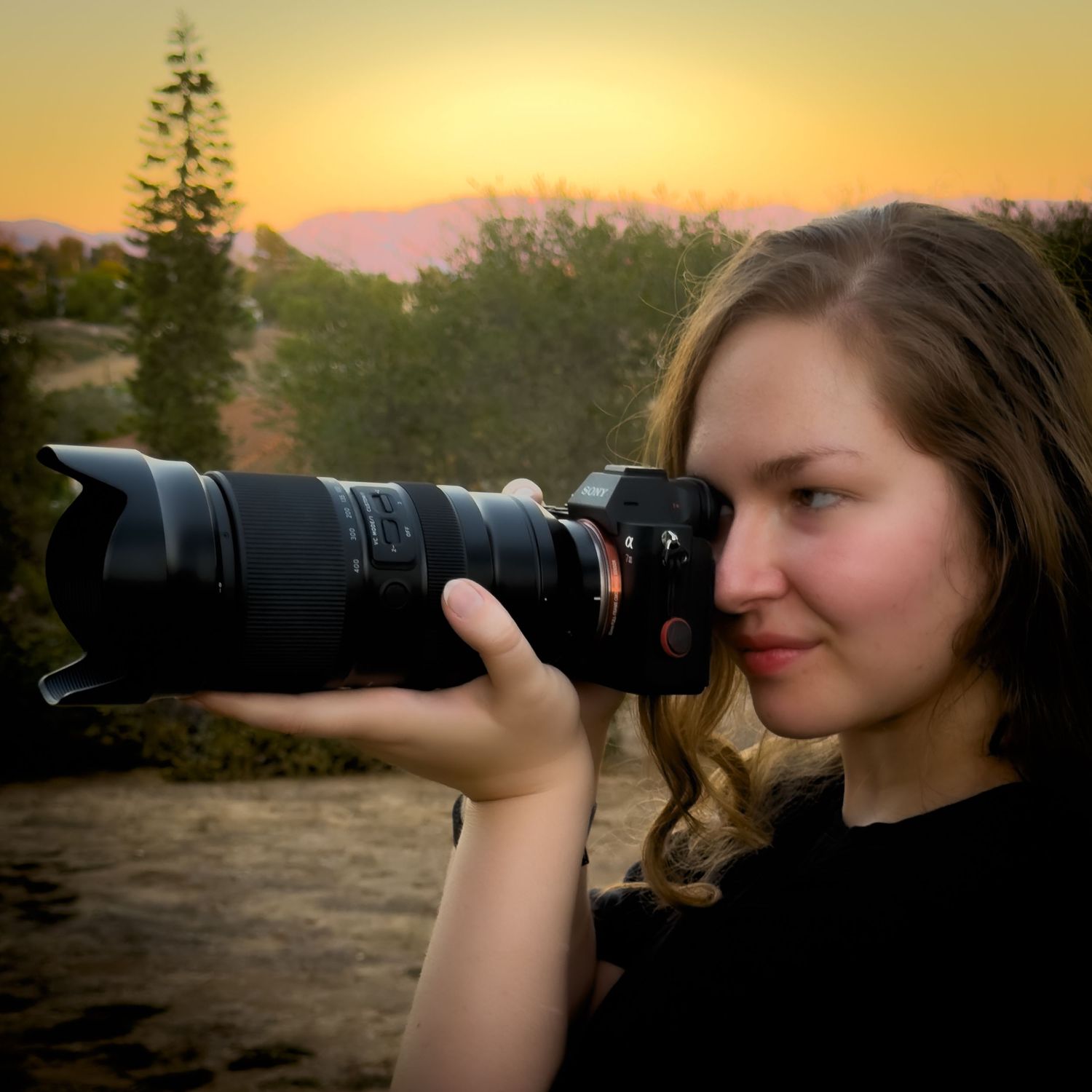 Young woman photographing with a professional Sony camera at golden hour sunset with trees and mountains in background.