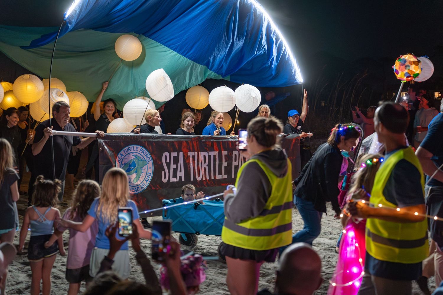 Sea Turtle Patrol volunteers carry glowing lanterns under a large illuminated canopy as families watch during the Hilton Head Lantern Parade