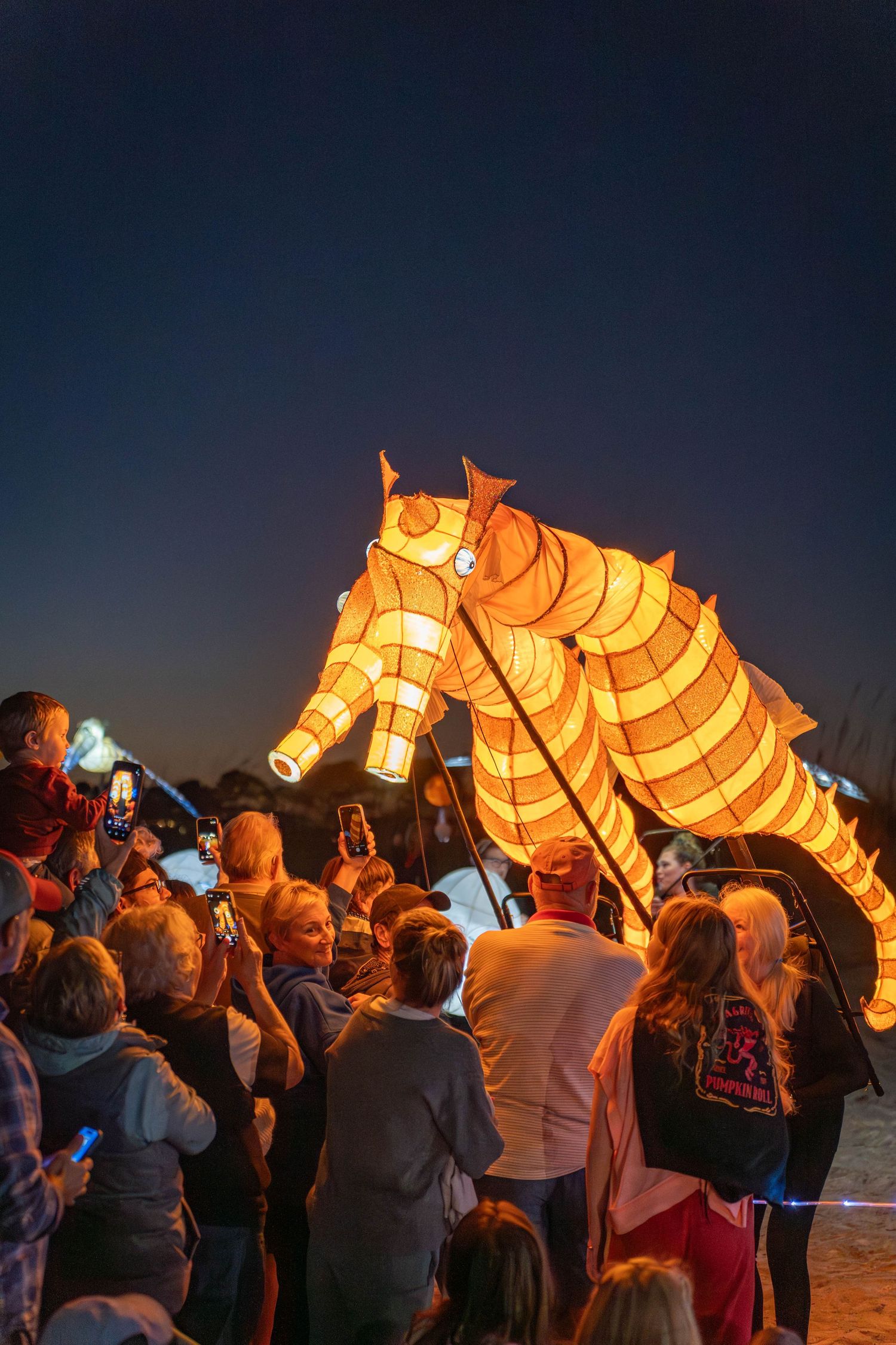 Two illuminated seahorse lanterns carried by performers as children and families watch during the beach Lantern Parade.