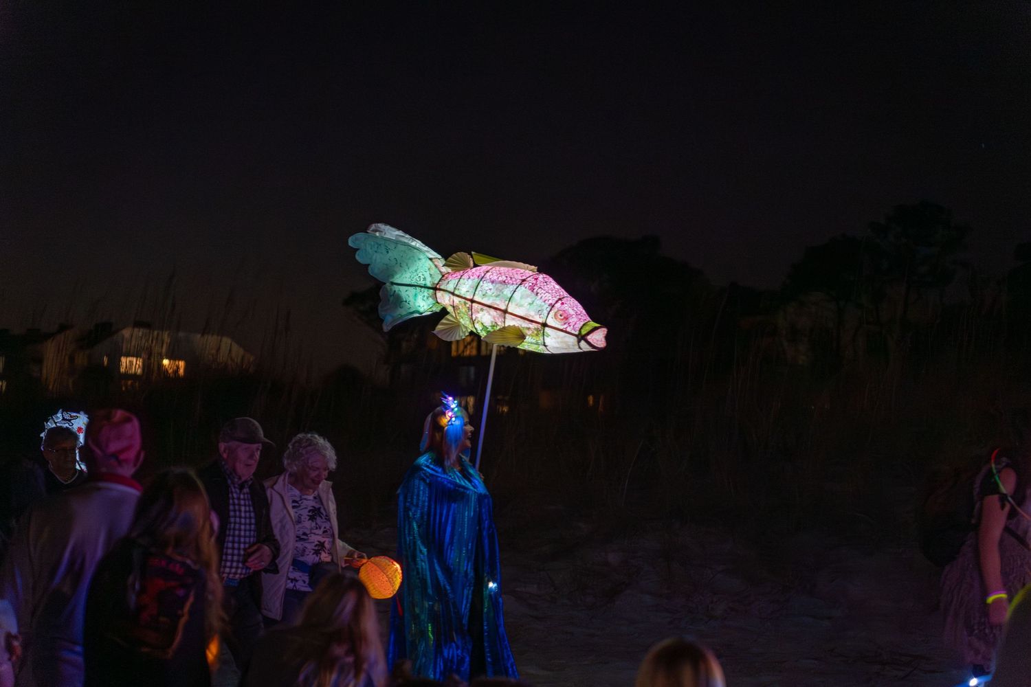 A performer dressed in blue carries a glowing pink and green fish lantern while walking past the crowd at the Hilton Head Lantern Parade.