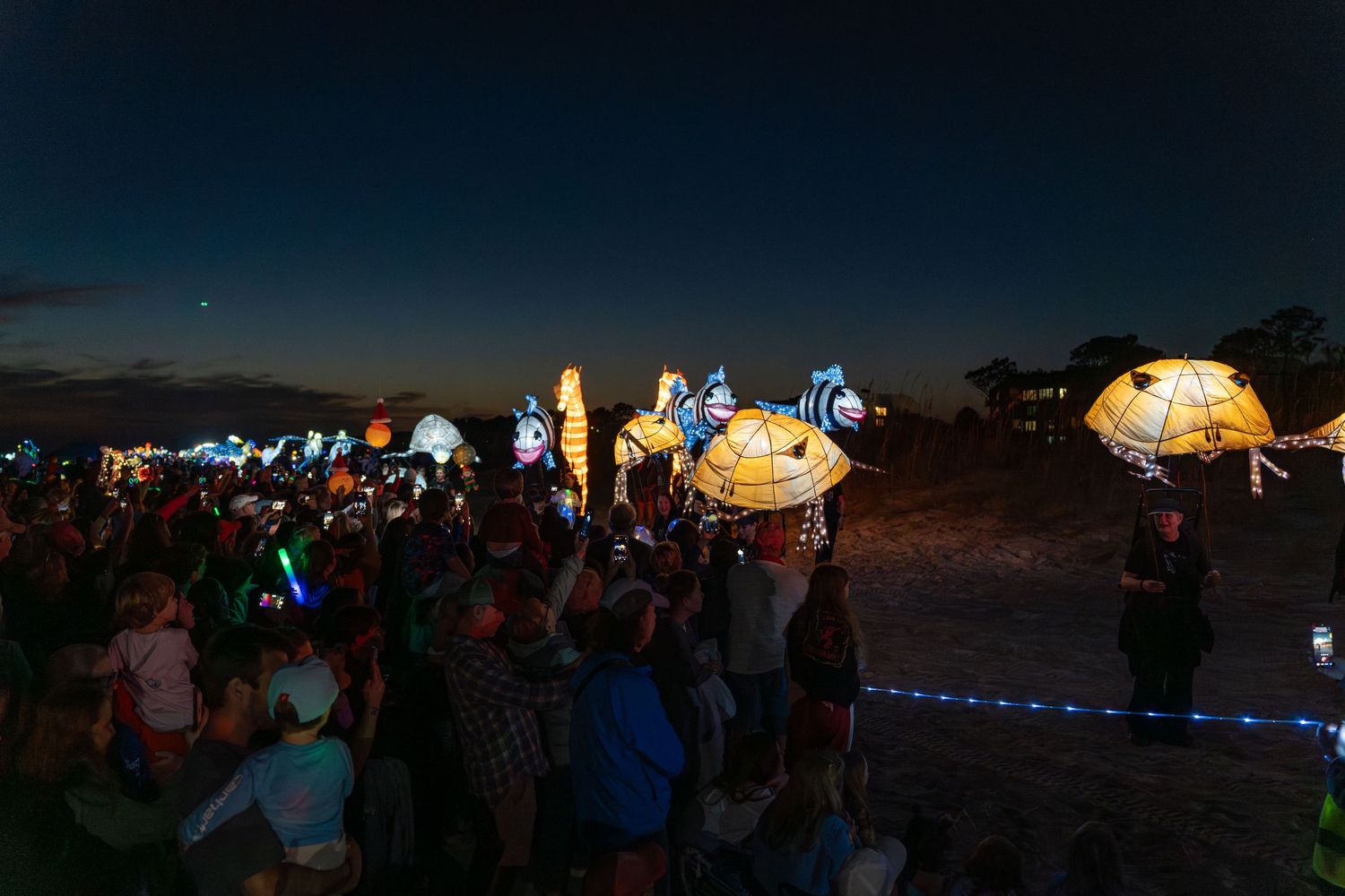 Crowd watching glowing sea-themed lantern puppets at sunset on Hilton Head Island beach during the Lantern Parade.