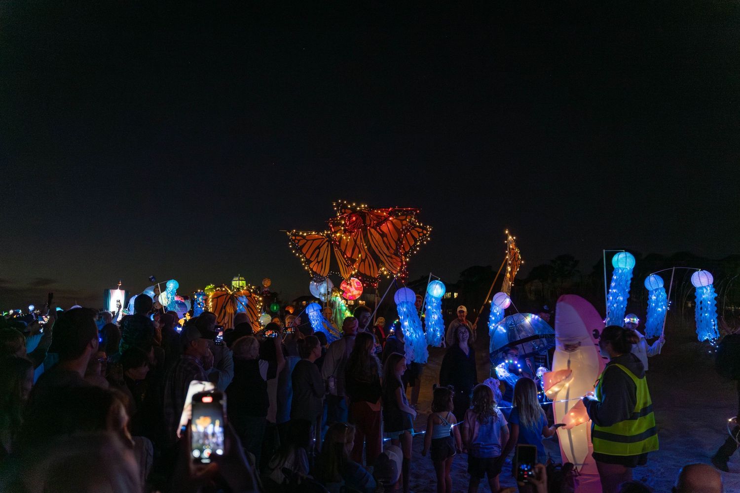 Giant glowing monarch butterfly lanterns shine above crowds as blue jellyfish and sea lanterns light the beach during the Hilton Head Parade