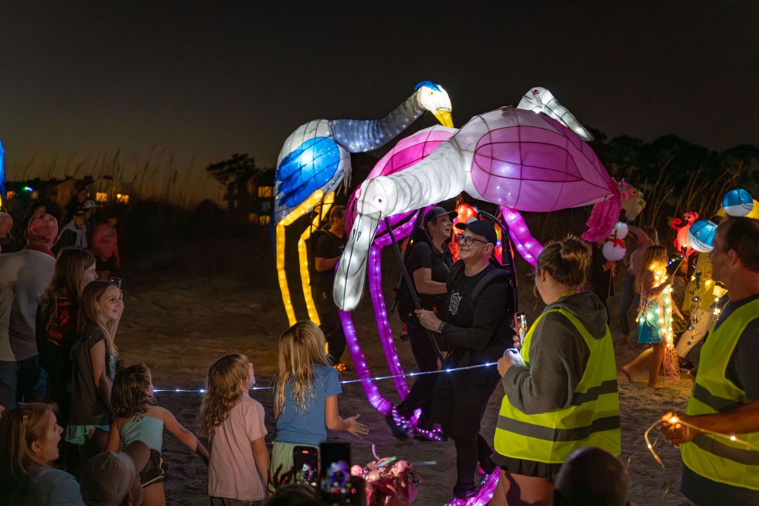 Colorful illuminated bird lanterns in bright pink, blue, and yellow walking among smiling spectators at the Hilton Head Lantern Parade.