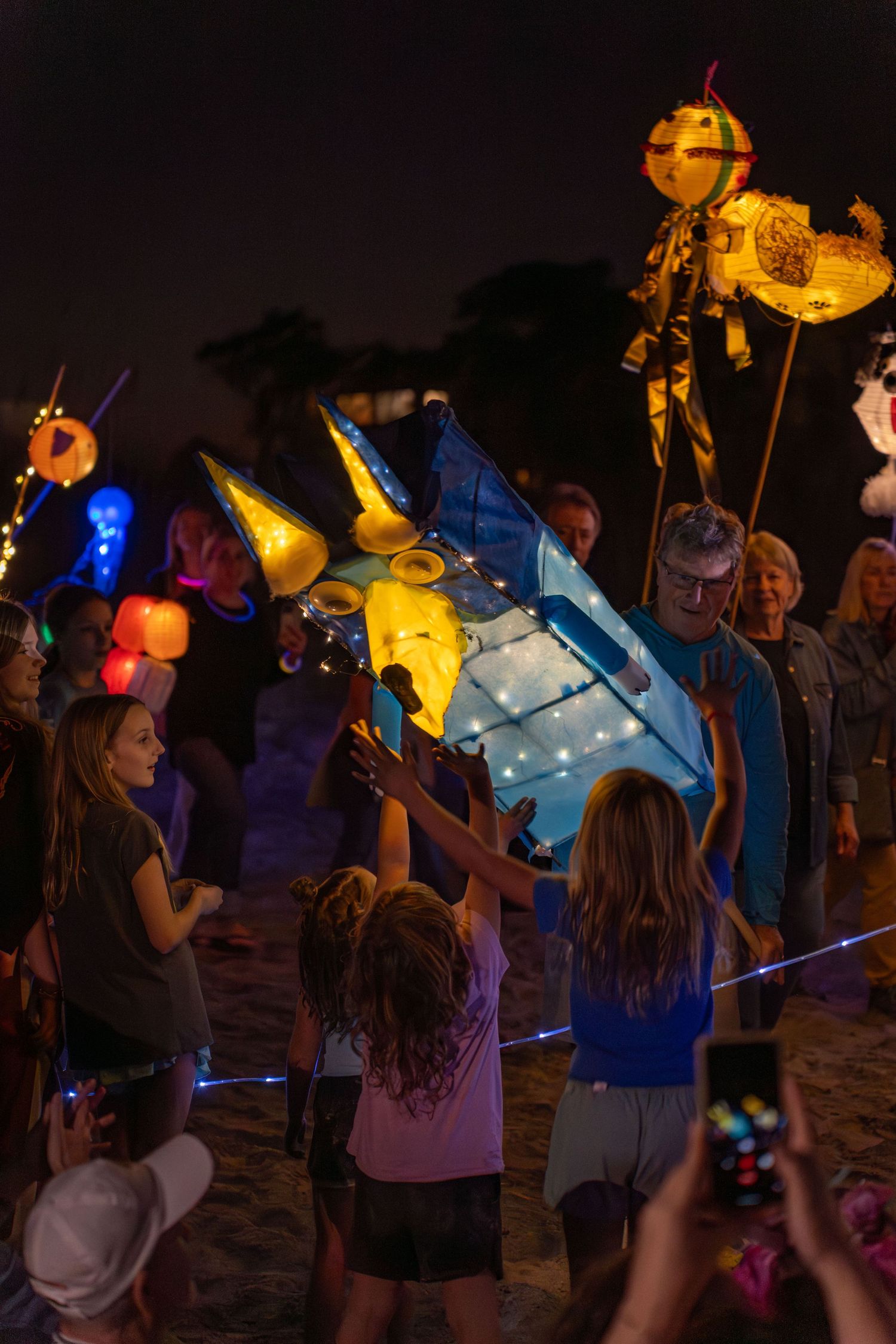 Children reaching toward a glowing bluey lantern creature during the Hilton Head Island Lantern Parade on the beach at night.