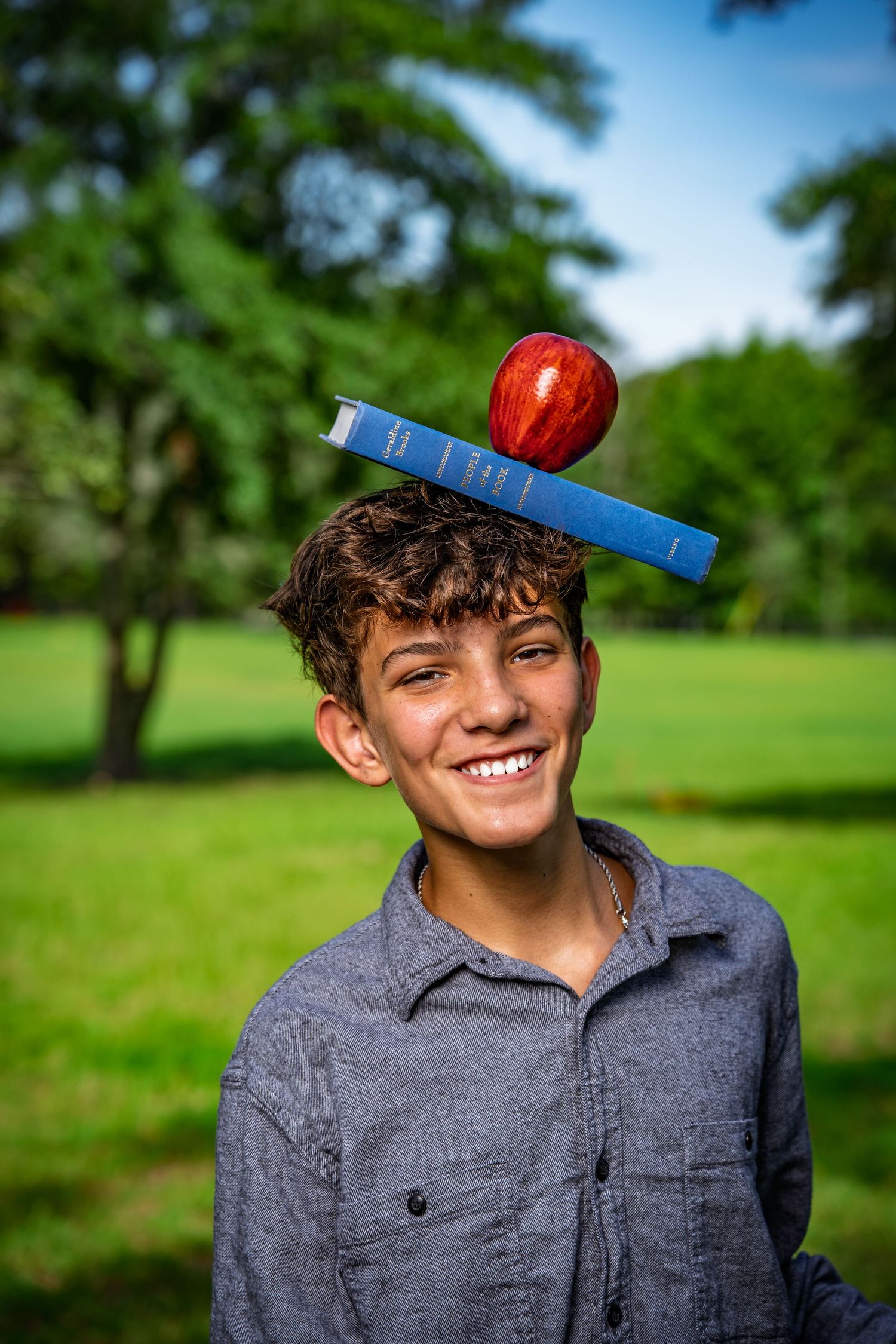 Smiling boy balancing a book and apple on his head during a back-to-school photo session at Red Barn Park in Bluffton, SC.