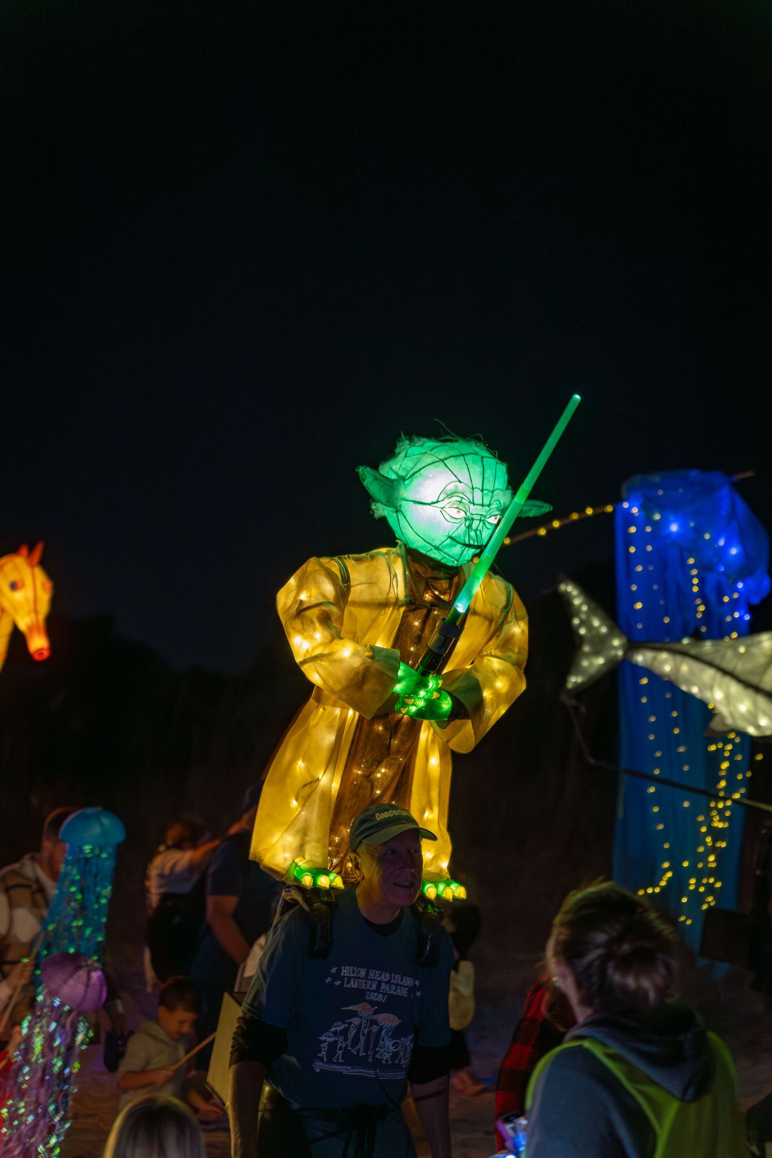 A large illuminated Yoda lantern with a bright green lightsaber appears among ocean-themed lanterns during the Hilton Head Lantern Parade.