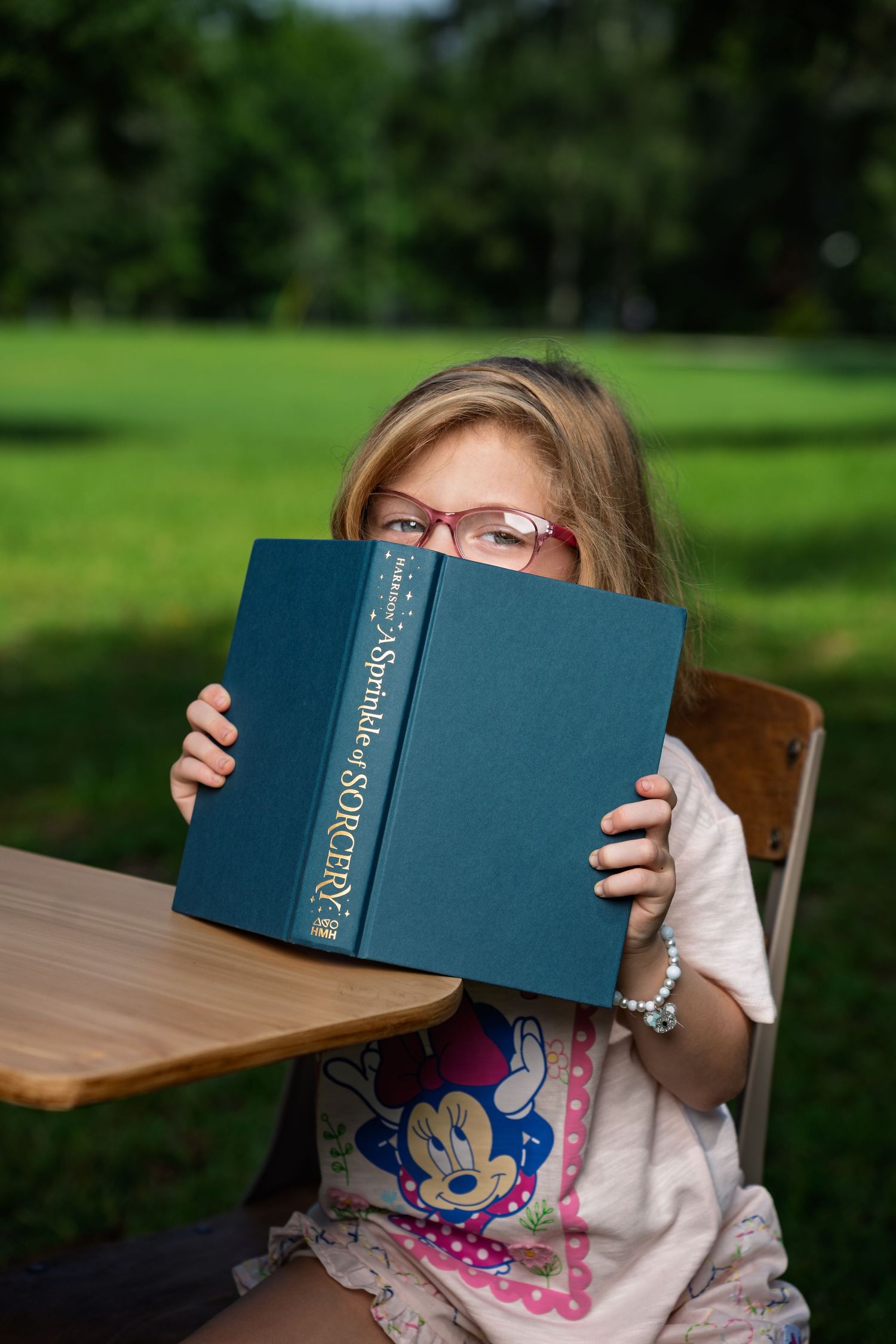 Young girl with glasses peeking over a book during a back-to-school photo session at Red Barn Park at New Riverside in Bluffton, SC.