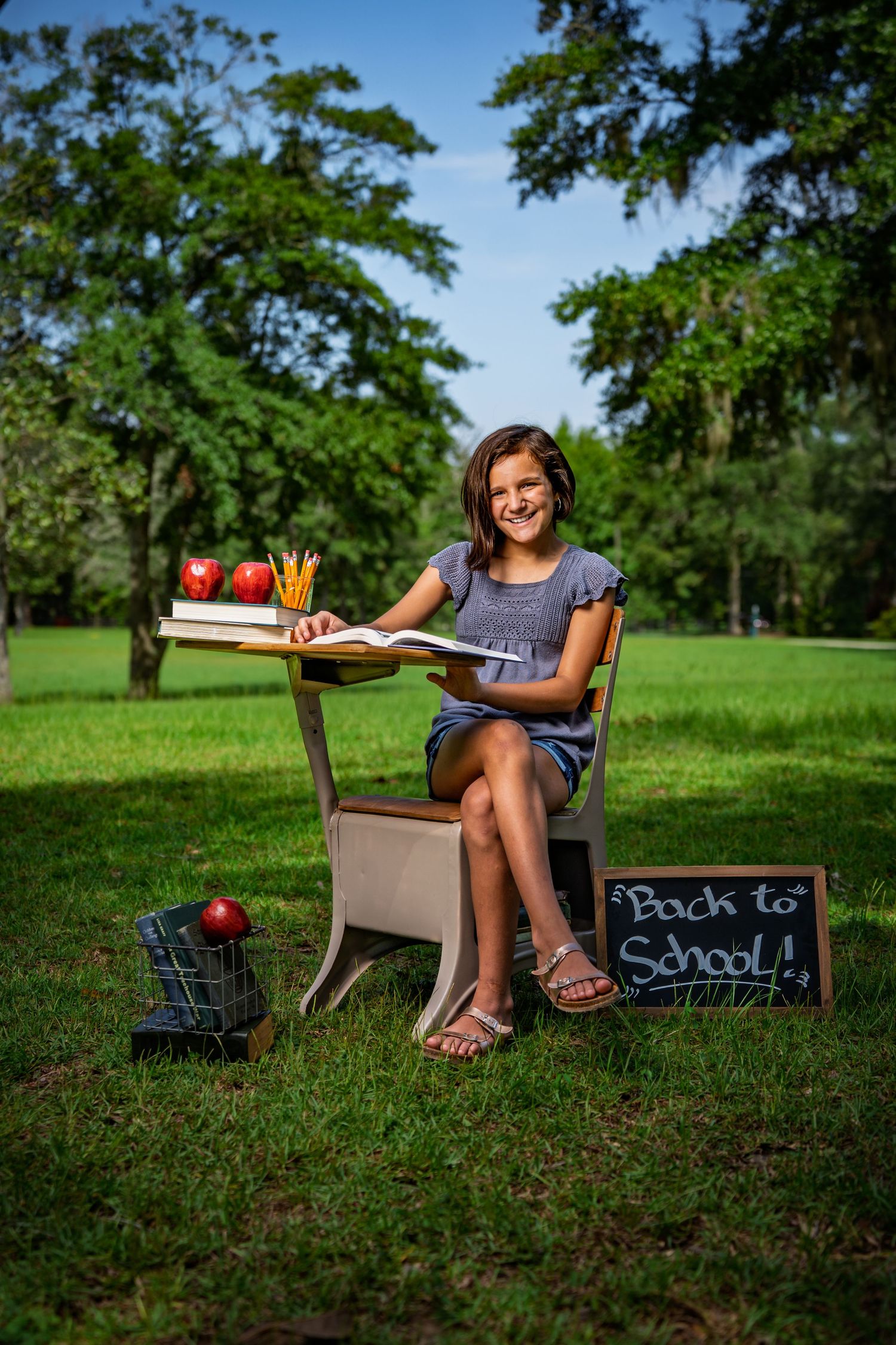 Girl smiling at a school desk with books and apples during a back-to-school photo session at Red Barn Park at New Riverside in Bluffton, SC.