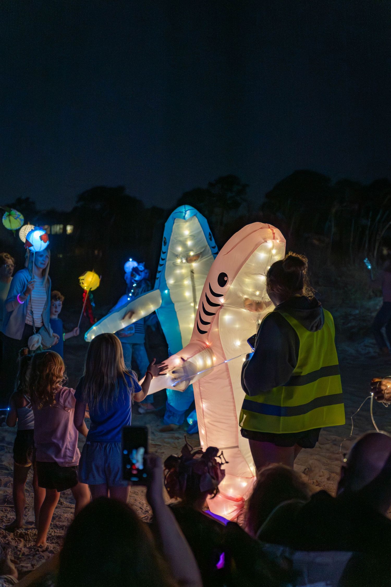 Children greet glowing shark lantern costumes as families walk the beach path during the nighttime Hilton Head Island Lantern Parade.