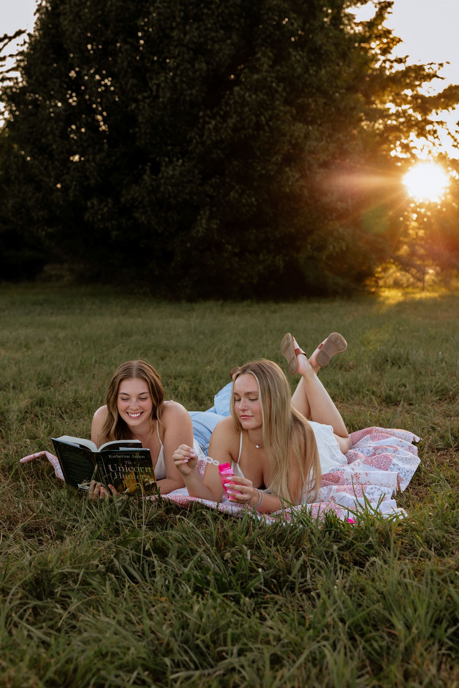 Two young women lying on a blanket in a field at sunset, reading a book and relaxing outdoors.