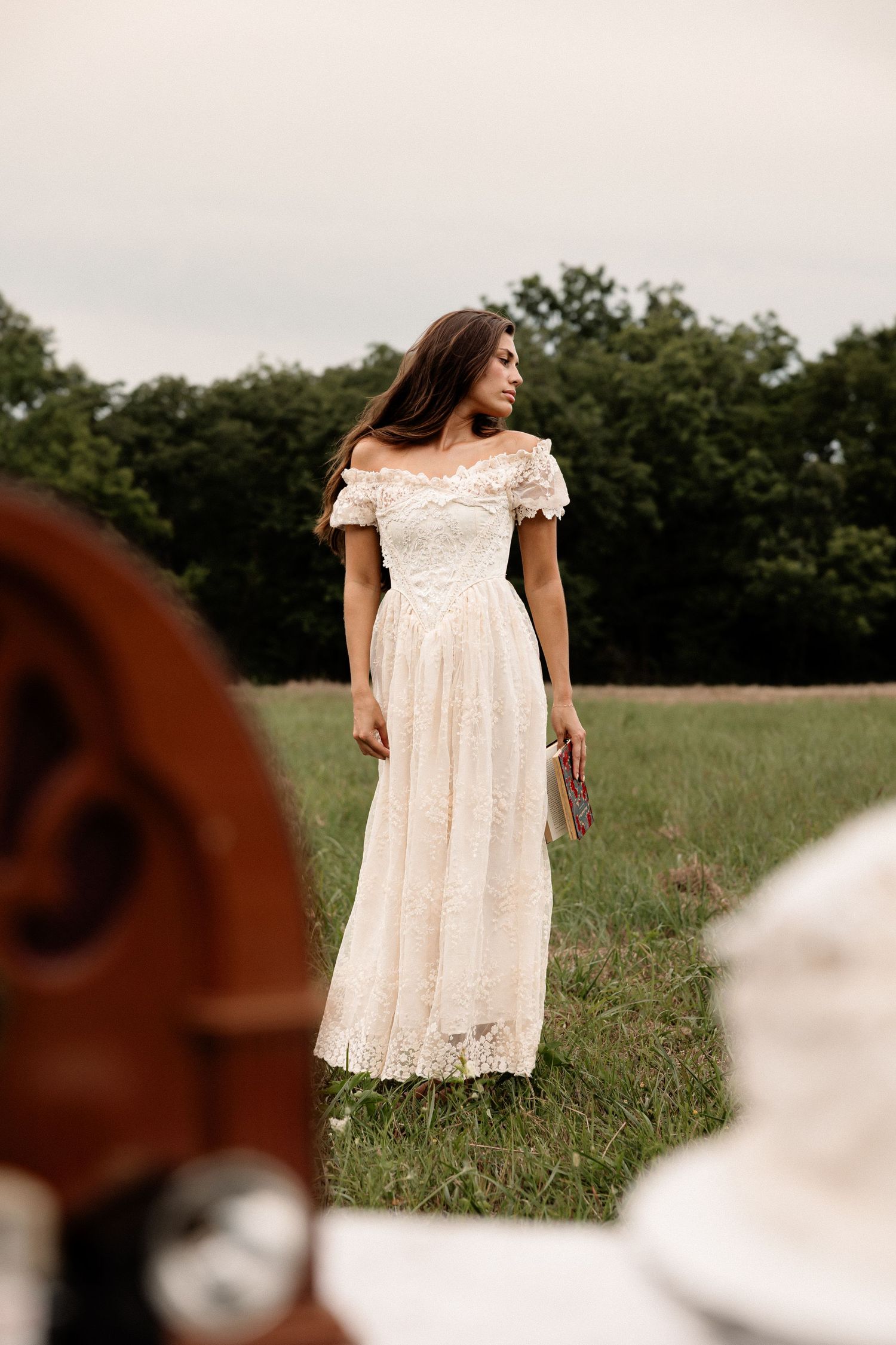 Woman in elegant off-shoulder ivory lace maxi dress standing in green field with trees in background.